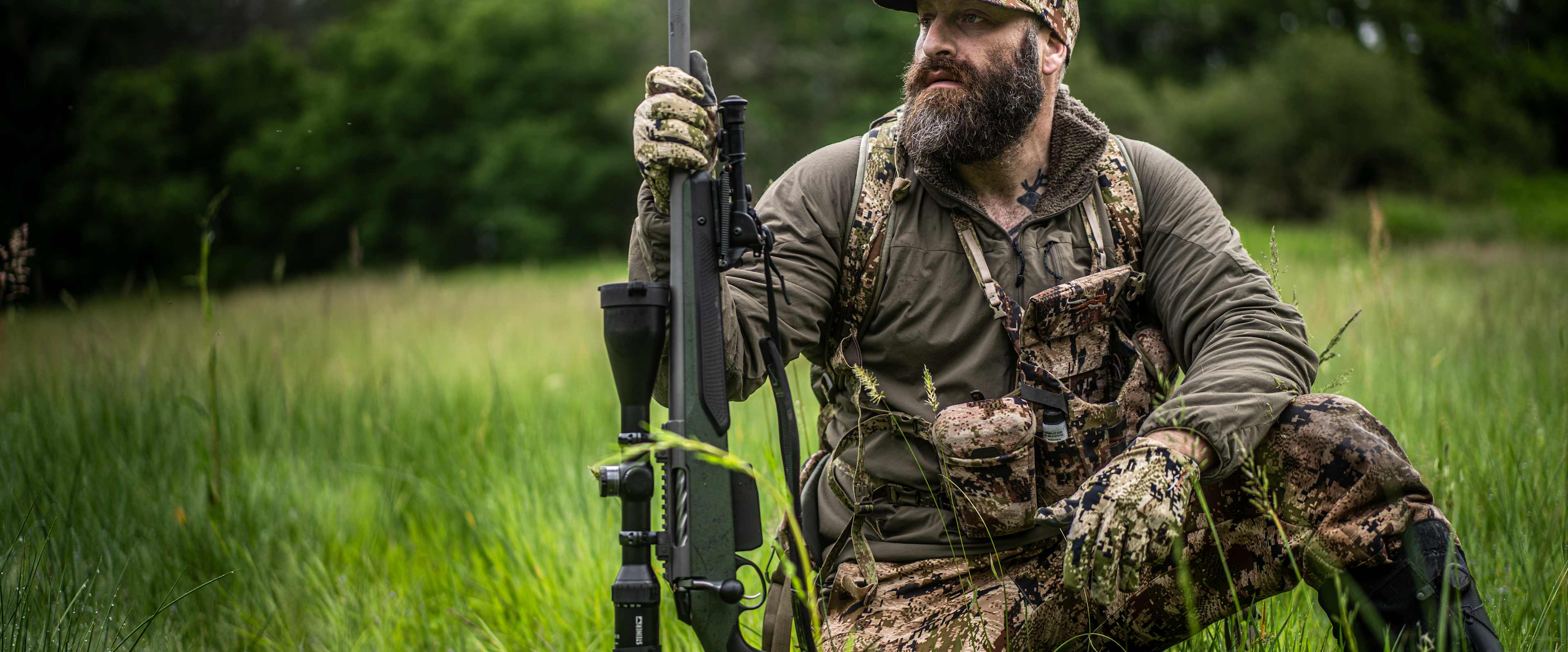 A bearded man in camouflage clothing and a cap sits in a grassy field, holding a rifle and surrounded by lush greenery.
