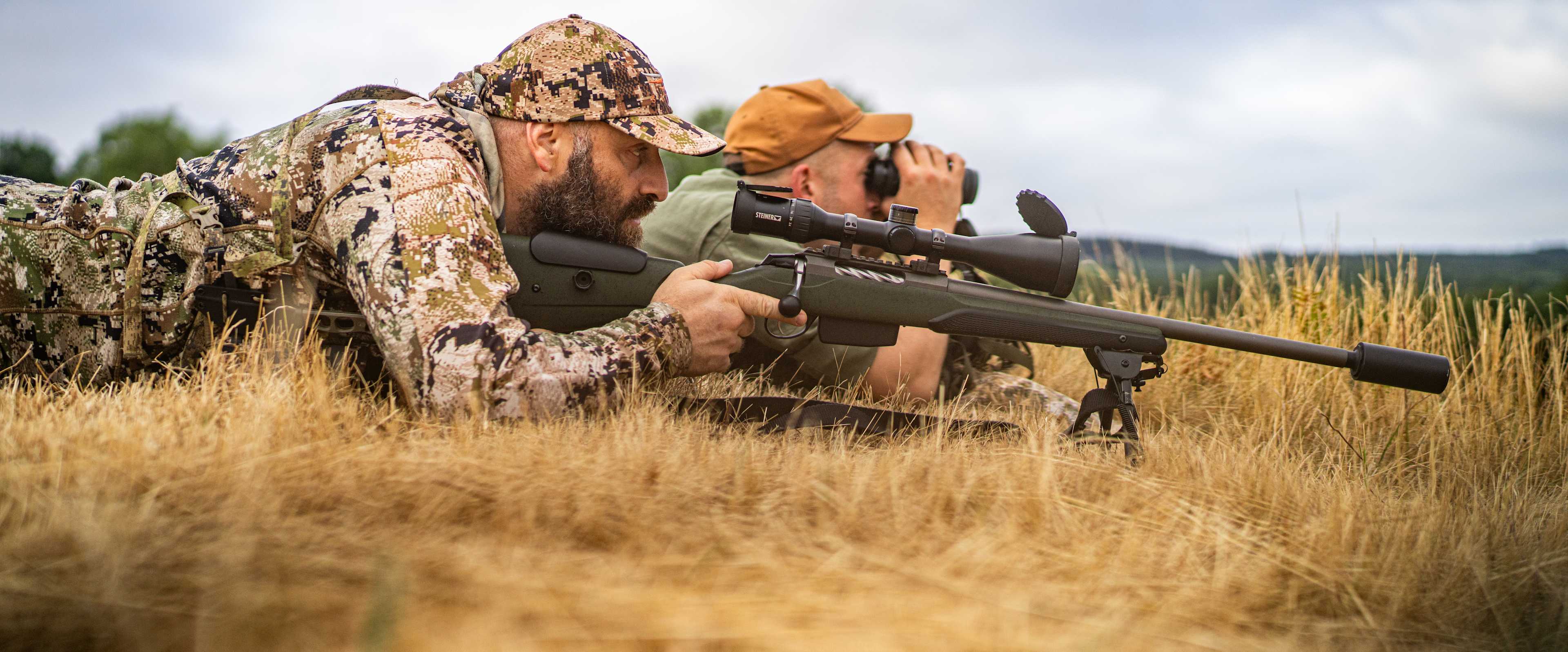 A camouflaged individual lies prone in a grassy field, aiming a rifle through the scope while the sky and clouds provide a backdrop.