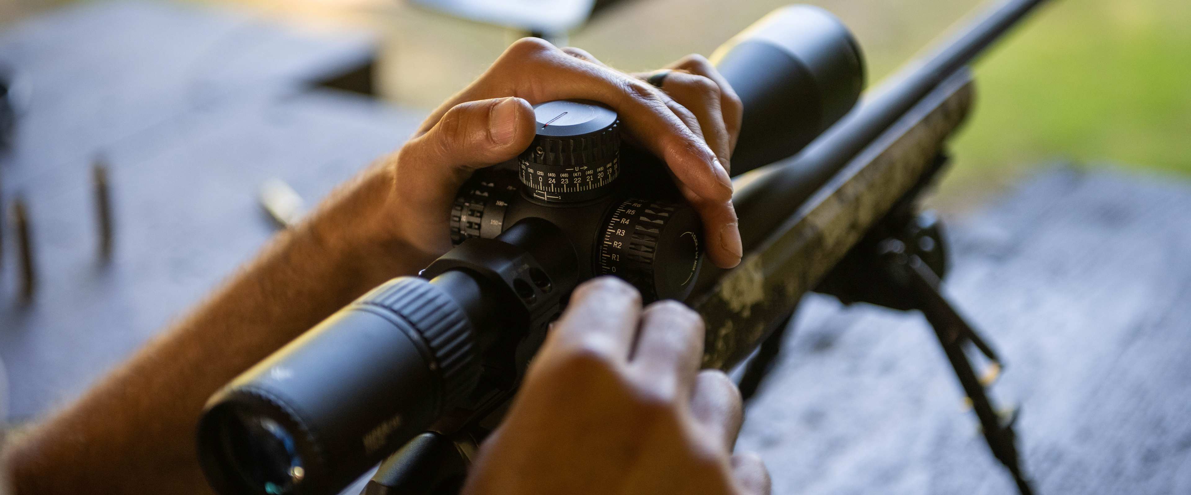 Hands adjust the scope on a rifle resting on a bipod, with a blurred outdoor setting in the background.