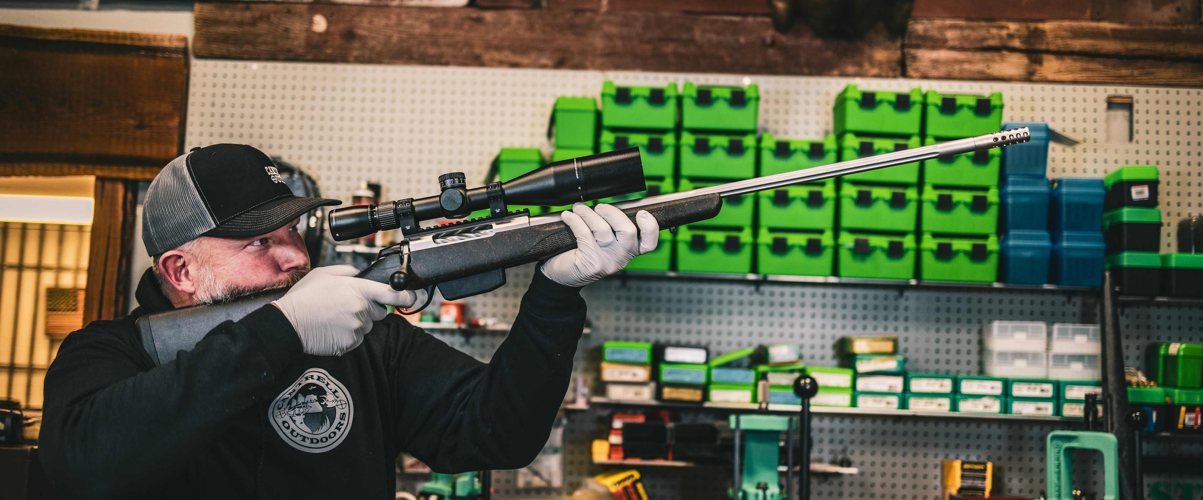 A person in a baseball cap is aiming a rifle at a wall of green targets in what appears to be a gun shop or shooting range.