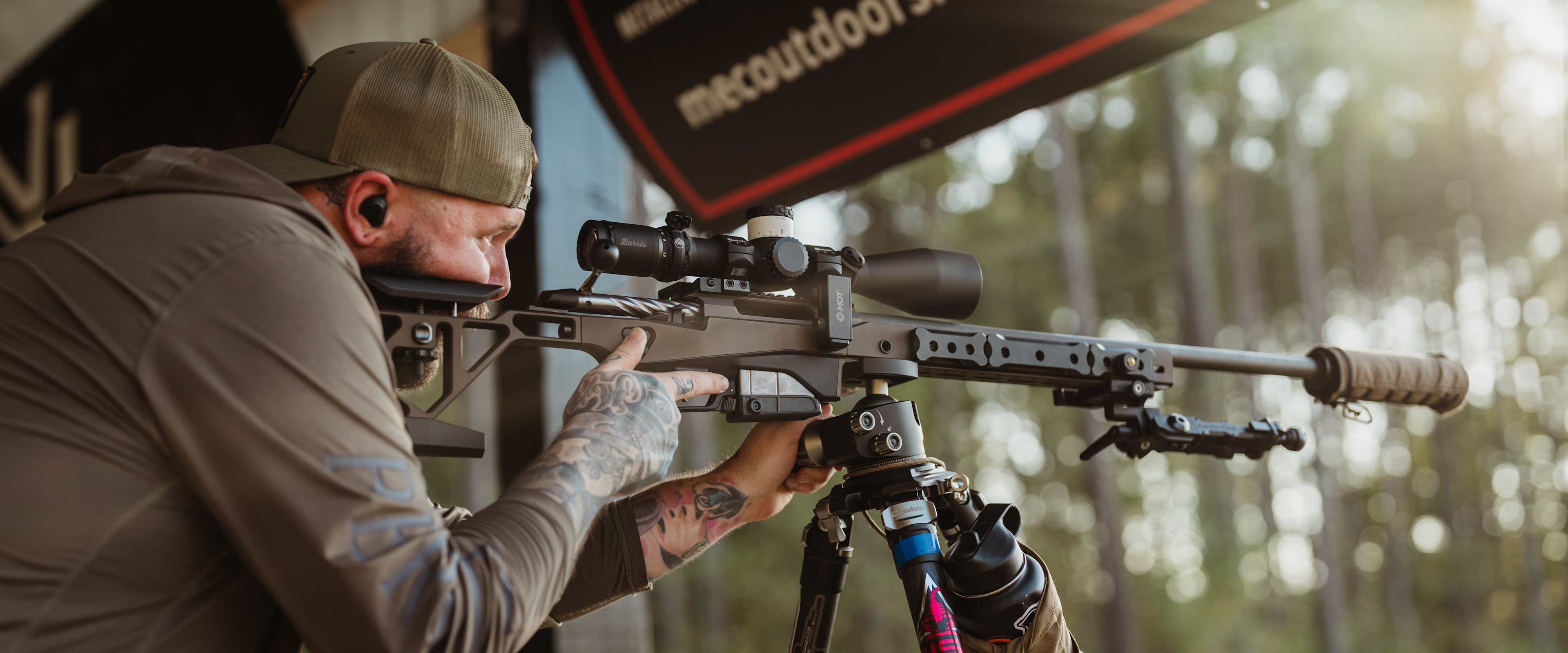 A person in camouflage clothing is aiming a rifle with a scope at a target, with a large advertisement banner visible in the background.
