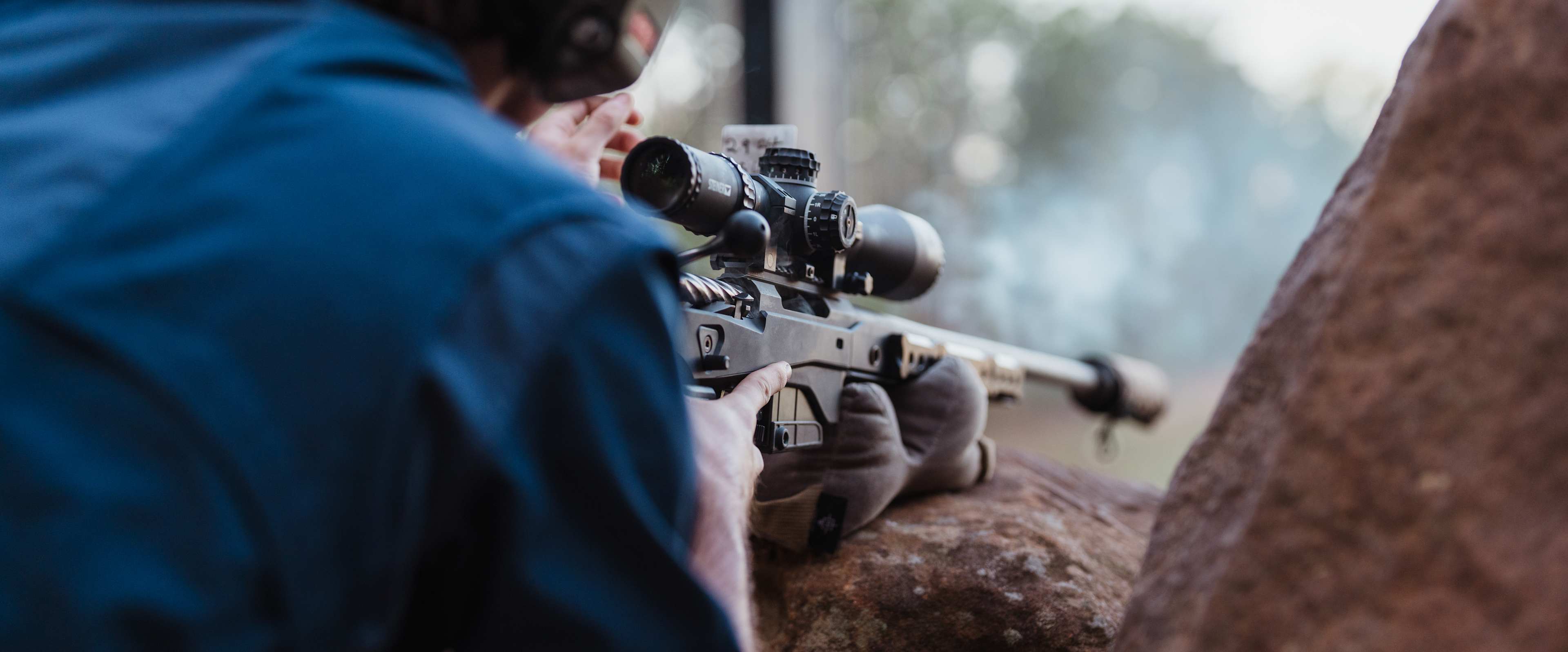 A person in a blue jacket is aiming a rifle with a scope towards the background, which appears to be a forested area.