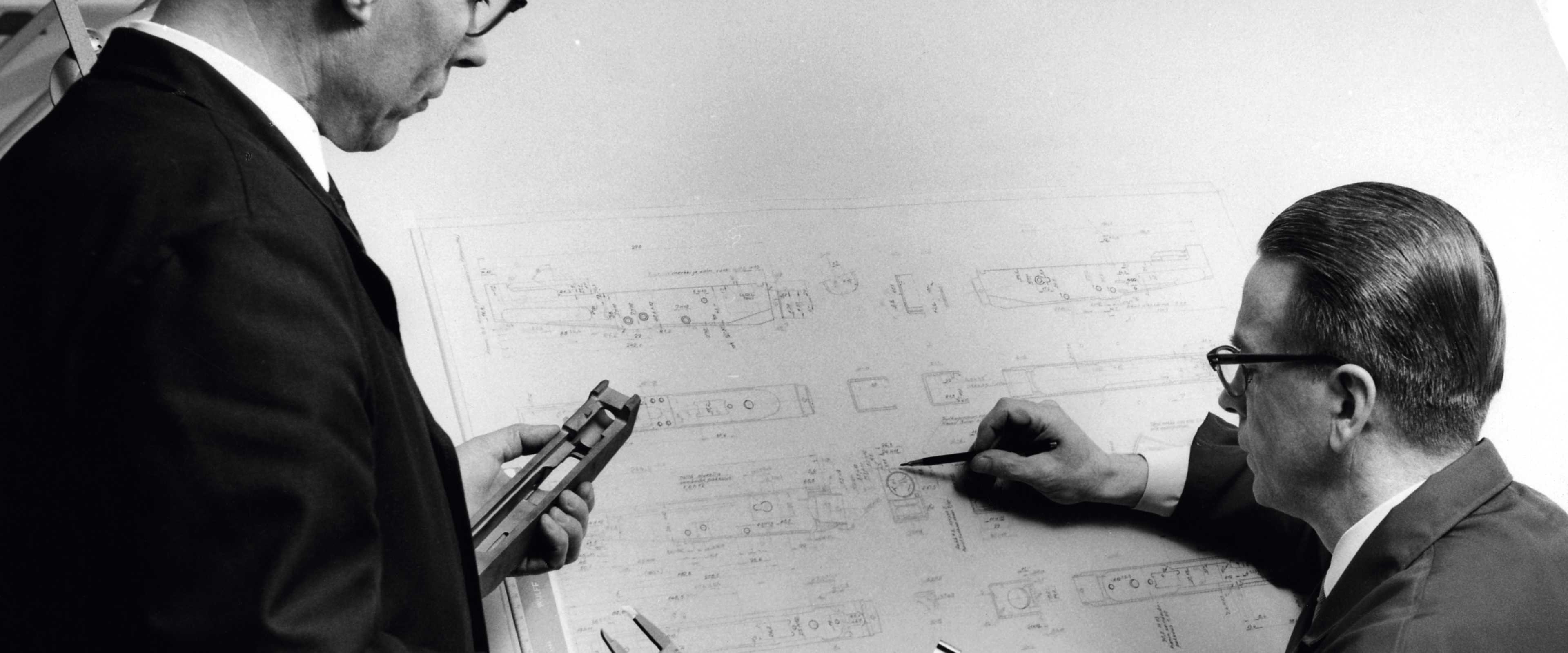 Two men in suits are standing in front of a whiteboard, engaged in discussion and examining the contents of the whiteboard.