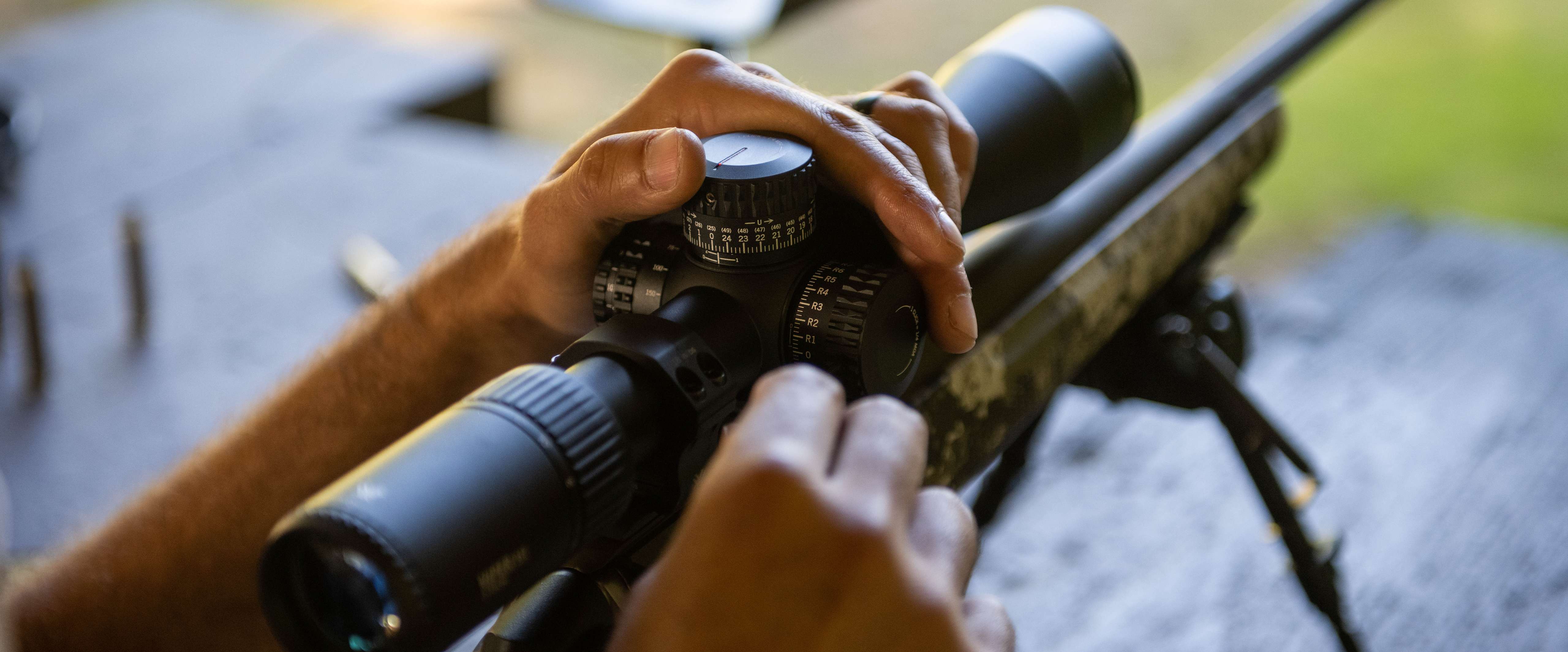 Hands adjust the scope on a rifle resting on a bipod, with a blurred outdoor setting in the background.