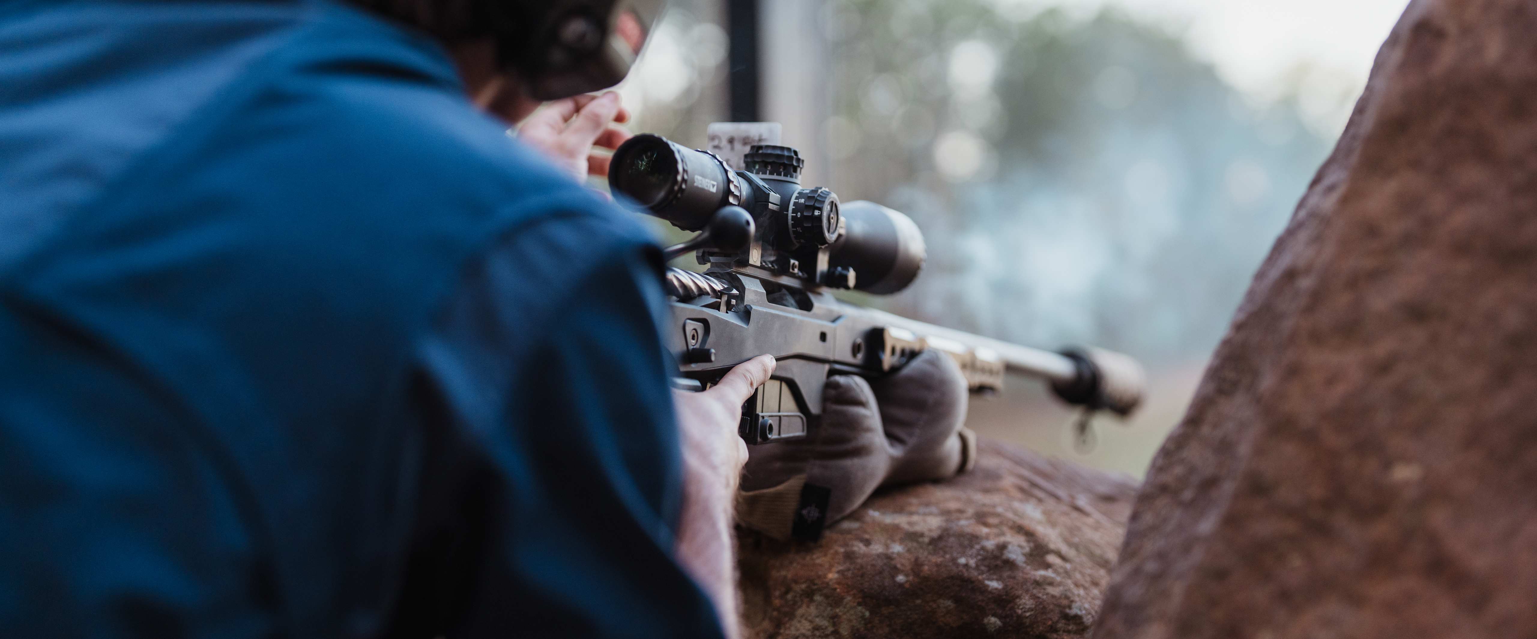 A person in a blue jacket is aiming a rifle with a scope towards the background, which appears to be a forested area.